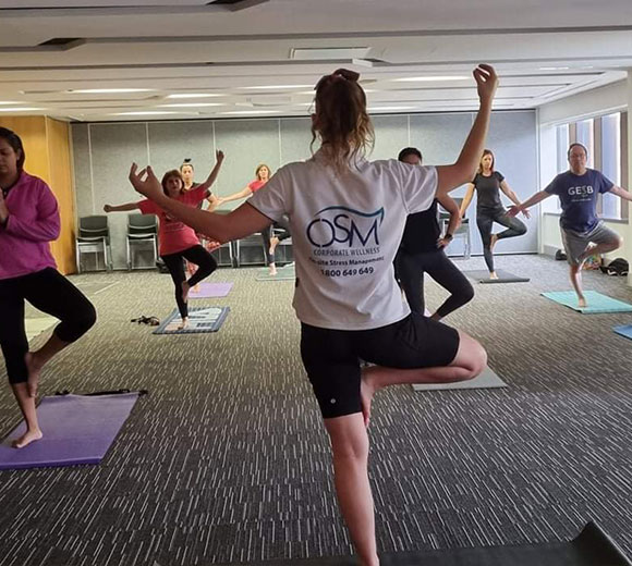 Corporate wellness yoga class in session, with employees practicing tree pose guided by an OSM instructor in a modern office space.